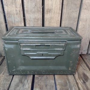 May include: An olive drab metal ammunition box with a hinged lid and a secure latch. The box has a rectangular shape and is labeled "AMMO BOX CAL .50 M2" along the bottom. The box is set against a wooden background.