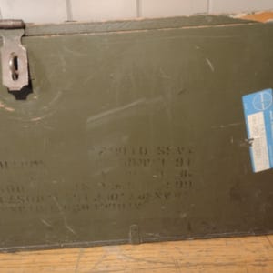 May include: A green wooden box with a metal latch and a blue label. The box has writing on it, including "2 W110 S21" and "1991".