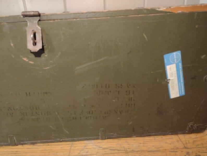 May include: A green wooden box with a metal latch and a blue label. The box has writing on it, including "2 W110 S21" and "1991".