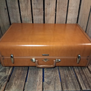 May include: A rectangular tan suitcase with a smooth finish, featuring a central handle, a central locking mechanism, and two silver-toned metal latches on the front. The suitcase has a structured design with rounded edges and a small brand logo.
