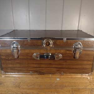 May include: A brown wooden storage trunk with metal accents and two latches. The trunk has a black handle and a metal ring on the top.