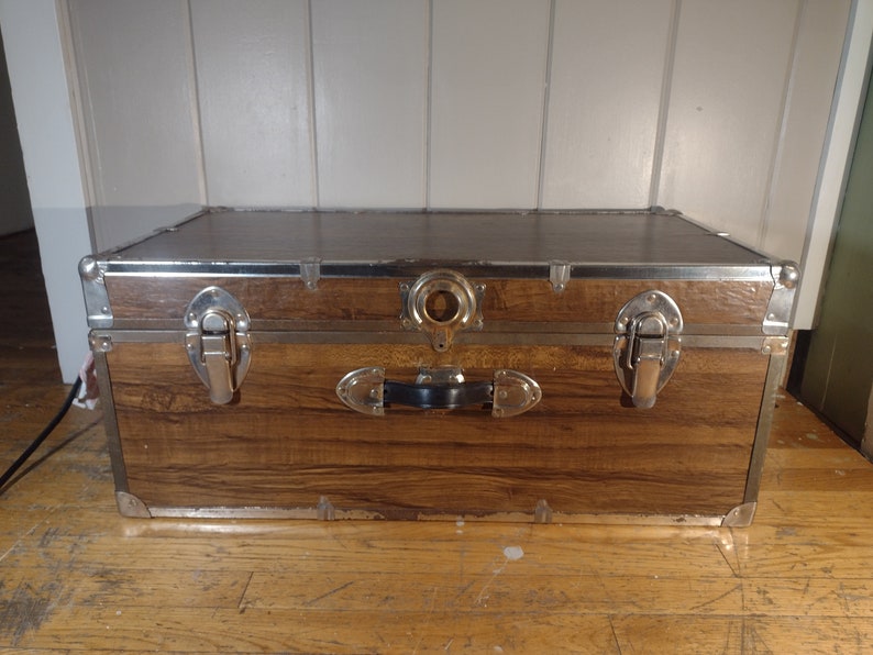 May include: A brown wooden storage trunk with metal accents and two latches. The trunk has a black handle and a metal ring on the top.