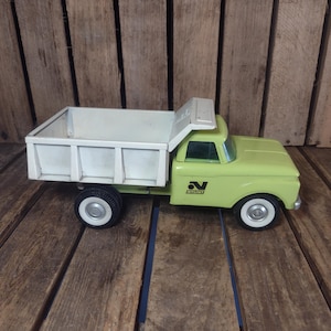 May include: A vintage lime green and white metal dump truck toy featuring a tilting cargo bed. The side of the truck cab displays the black Nylint logo. It has black plastic wheels with white rims and a clear plastic windshield on the cab.