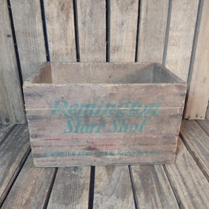 May include: A weathered wooden crate with the words "Remington Shur Shot" in green and red lettering. The crate is made of wooden planks and sits on a wooden surface. The crate is empty.