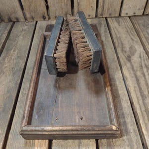 May include: An antique wooden shoe shine kit. The kit includes a rectangular wooden tray with a raised edge and two shoe brushes with dark metal backs and brown bristles. The wood has a weathered appearance.