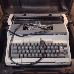 May include: A vintage Corona Super Correct XT typewriter in its black carrying case. The typewriter has a white and gray body with a full QWERTY keyboard. The text "CORONA" and "SUPER CORRECT XT" are visible on the machine.
