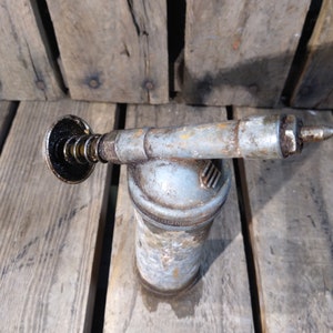 May include: A vintage metal grease gun with a black plastic nozzle and a silver metal body. The gun is sitting on a wooden surface.