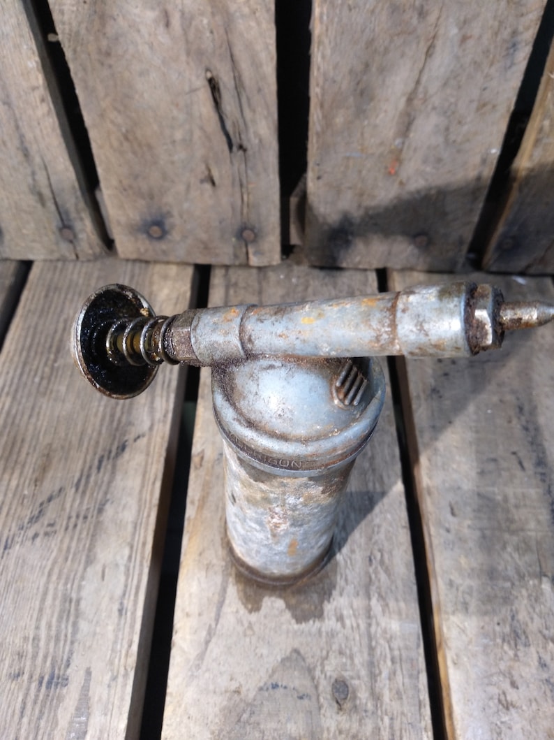 May include: A vintage metal grease gun with a black plastic nozzle and a silver metal body. The gun is sitting on a wooden surface.