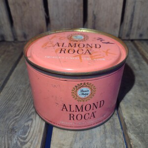 May include: A pink metal tin with a gold lid. The lid has the text "Almond Roca" and "America's Finest Confection" printed on it. The tin has a gold label with the text "Almond Roca" and "America's Finest Confection Made by Brown & Haley" printed on it.