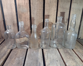 May include: Seven clear glass bottles of various shapes and sizes, arranged in a row on a wooden surface. The bottles are empty and appear to be vintage.