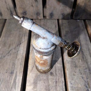 May include: A rusty, gray metal grease gun with a black plastic nozzle and a spring-loaded plunger. The gun is sitting on a wooden surface.