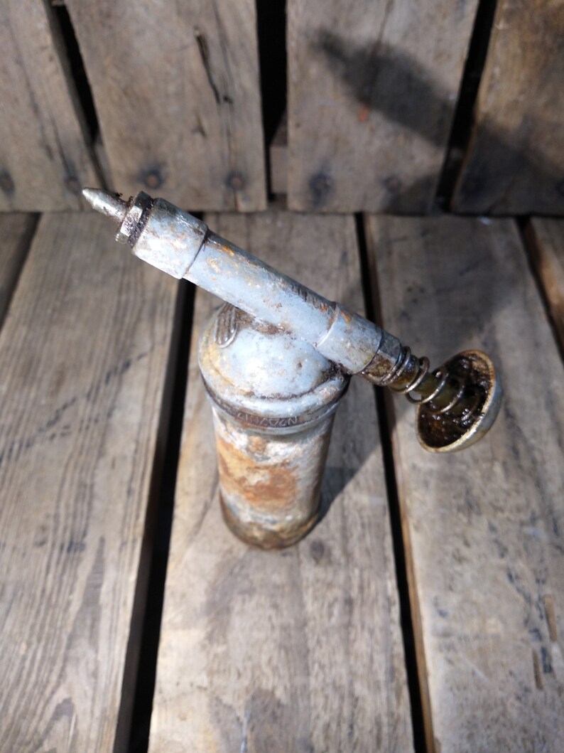 May include: A rusty, gray metal grease gun with a black plastic nozzle and a spring-loaded plunger. The gun is sitting on a wooden surface.