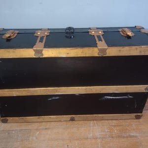 Puede incluir: Un ba&uacute;l vintage negro con ribetes dorados y herrajes de lat&oacute;n. El ba&uacute;l tiene dos secciones y est&aacute; hecho de madera.
