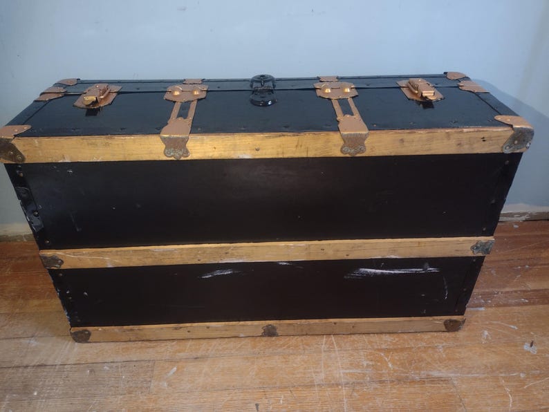 Puede incluir: Un ba&uacute;l vintage negro con ribetes dorados y herrajes de lat&oacute;n. El ba&uacute;l tiene dos secciones y est&aacute; hecho de madera.