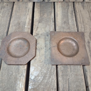 May include: Two copper-colored metal dishes. One is octagonal, the other square. Both have a circular indentation in the center. The dishes are displayed on a weathered wooden surface.