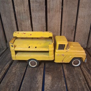 May include: A vintage yellow metal toy truck featuring a flatbed cargo area with side rails, a cabin with windows, and four black wheels with white rims. The paint shows signs of wear and chipping, revealing the metal surface underneath.