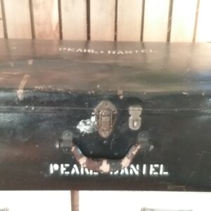 May include: Black metal vintage suitcase with two latches and a handle. The suitcase has the words "Pearl Daniel" written on the top and bottom.