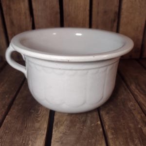 May include: A white ceramic chamber pot featuring a smooth, rounded basin with a wide, flared rim and a single side handle. The exterior surface is decorated with a subtle, embossed scalloped pattern that encircles the midsection of the vessel.
