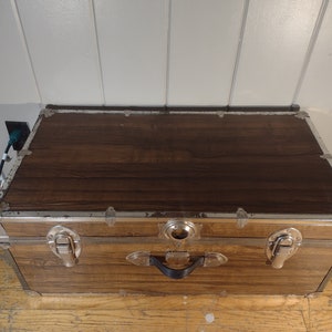 May include: A vintage wooden storage trunk with metal accents and a black handle. The trunk has a brown woodgrain finish and silver metal trim.