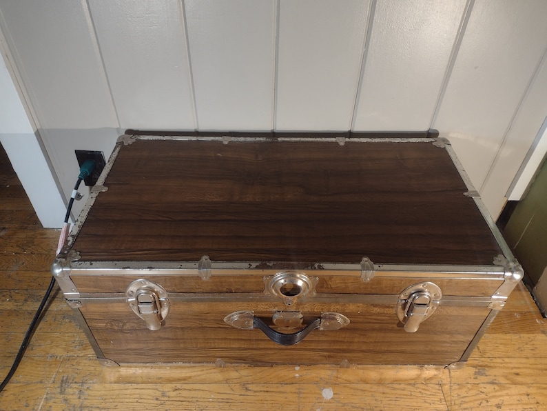 May include: A vintage wooden storage trunk with metal accents and a black handle. The trunk has a brown woodgrain finish and silver metal trim.