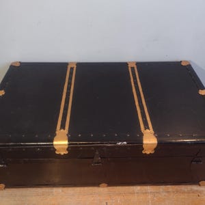 Puede incluir: Un ba&uacute;l vintage negro con detalles dorados y herrajes de lat&oacute;n. El ba&uacute;l tiene una tapa con bisagras y una cerradura.