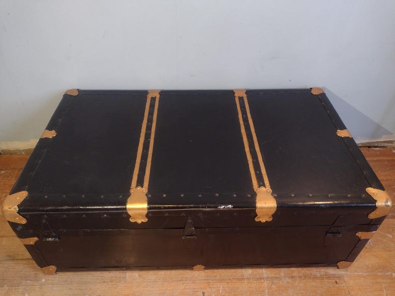Puede incluir: Un ba&uacute;l vintage negro con detalles dorados y herrajes de lat&oacute;n. El ba&uacute;l tiene una tapa con bisagras y una cerradura.