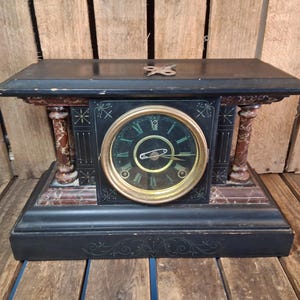 May include: A black mantel clock with a gold-toned circular face, Roman numerals, and two hands. The clock features red marble-patterned columns on each side, decorative etched line patterns, and a small metal winding key resting on the top surface.