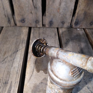 May include: A vintage, silver-colored oil can with a black, spring-loaded nozzle. The can has a worn, rusty appearance and the nozzle is filled with dark oil.