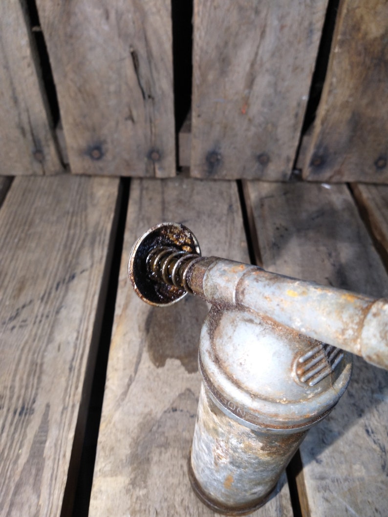 May include: A vintage, silver-colored oil can with a black, spring-loaded nozzle. The can has a worn, rusty appearance and the nozzle is filled with dark oil.
