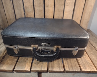 May include: A vintage black suitcase with silver trim and a black handle. The suitcase has a textured surface and metal clasps. The handle is made of a black, textured material. The suitcase is sitting on a wooden surface.