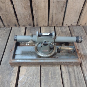 May include: A vintage transit level, a surveying instrument, rests on a wooden base. The level is painted gray with brass accents and features a bubble level and a circular protractor. The instrument is designed for measuring angles and elevations.