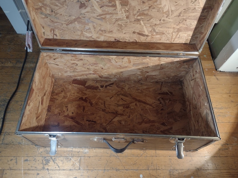 May include: An empty, open, wooden storage trunk with silver metal accents and a black handle. The inside of the trunk is lined with plywood.