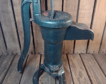 May include: An antique, dark teal cast iron water pump. The pump features a curved handle, a circular basin, and a spout. The pump is mounted on a U-shaped base. The background is a wooden surface.