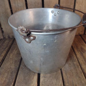May include: A vintage, aluminum bucket with a wire handle and wooden grip. The bucket has a slightly tapered shape and is set against a rustic wooden background. The metal shows signs of wear, suggesting age and use. The bucket is empty.