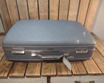 May include: A vintage blue suitcase with a rounded top and silver hardware. The suitcase has a handle and two latches. A small tag hangs from the handle. The suitcase is sitting on a wooden surface.