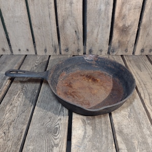 May include: A black cast iron skillet with a rusty interior. The skillet is sitting on a wooden surface.