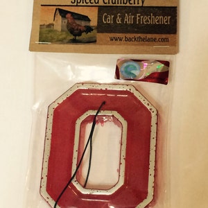 May include: A red car air freshener in the shape of the letter "O" with a white border. The packaging includes a label with the text "Back The Lane Creations Spiced Cranberry" and a picture of a rooster.