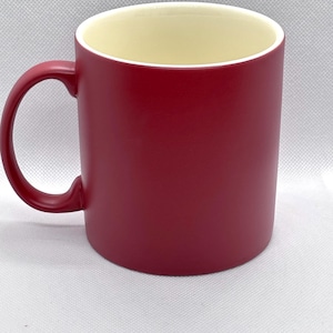 Puede incluir: Una taza cil&iacute;ndrica de cer&aacute;mica con un acabado exterior rojo oscuro mate y un interior liso de color crema. La taza cuenta con un asa roja a juego en forma de C en el lado izquierdo, ofreciendo un dise&ntilde;o sencillo y funcional para bebidas.