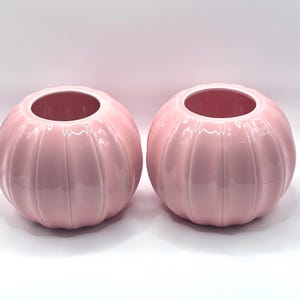 May include: Two matching spherical ceramic vases in a glossy light pink finish. Each vase features a distinct ribbed or fluted texture with vertical indentations running from the circular opening down to the base, creating a pumpkin-like shape.