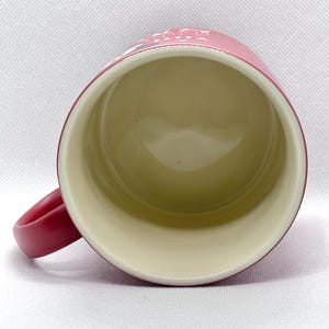 Puede incluir: Una vista desde arriba de una taza cil&iacute;ndrica con un exterior rojo mate y un interior de color crema liso. Un asa curva se extiende desde el lado izquierdo. Una parte del texto blanco es visible en el borde exterior superior de la taza roja.