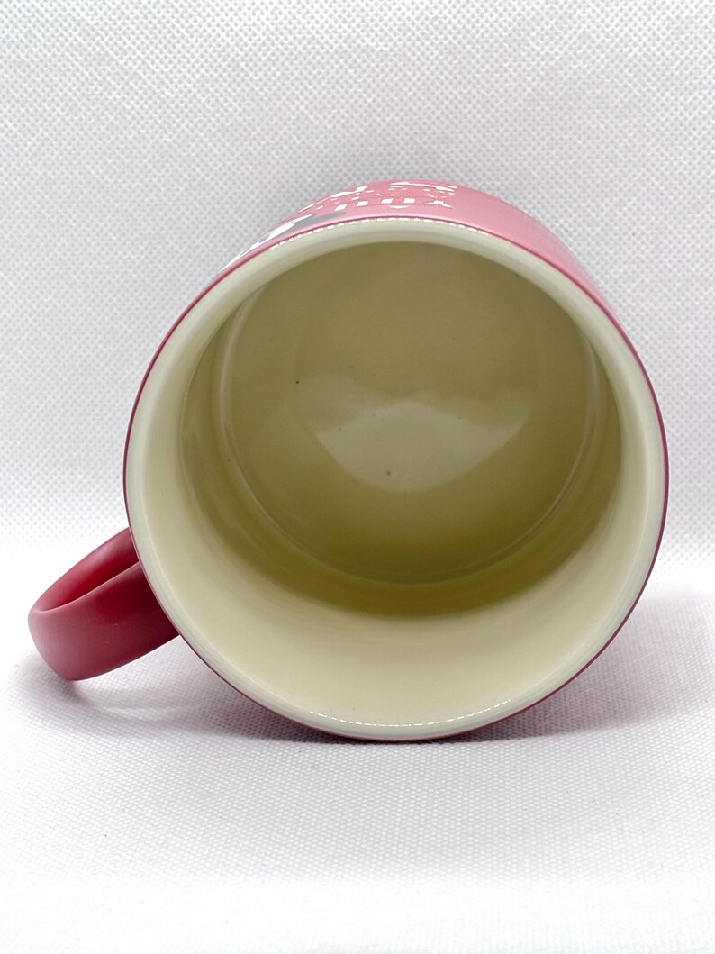 Puede incluir: Una vista desde arriba de una taza cil&iacute;ndrica con un exterior rojo mate y un interior de color crema liso. Un asa curva se extiende desde el lado izquierdo. Una parte del texto blanco es visible en el borde exterior superior de la taza roja.