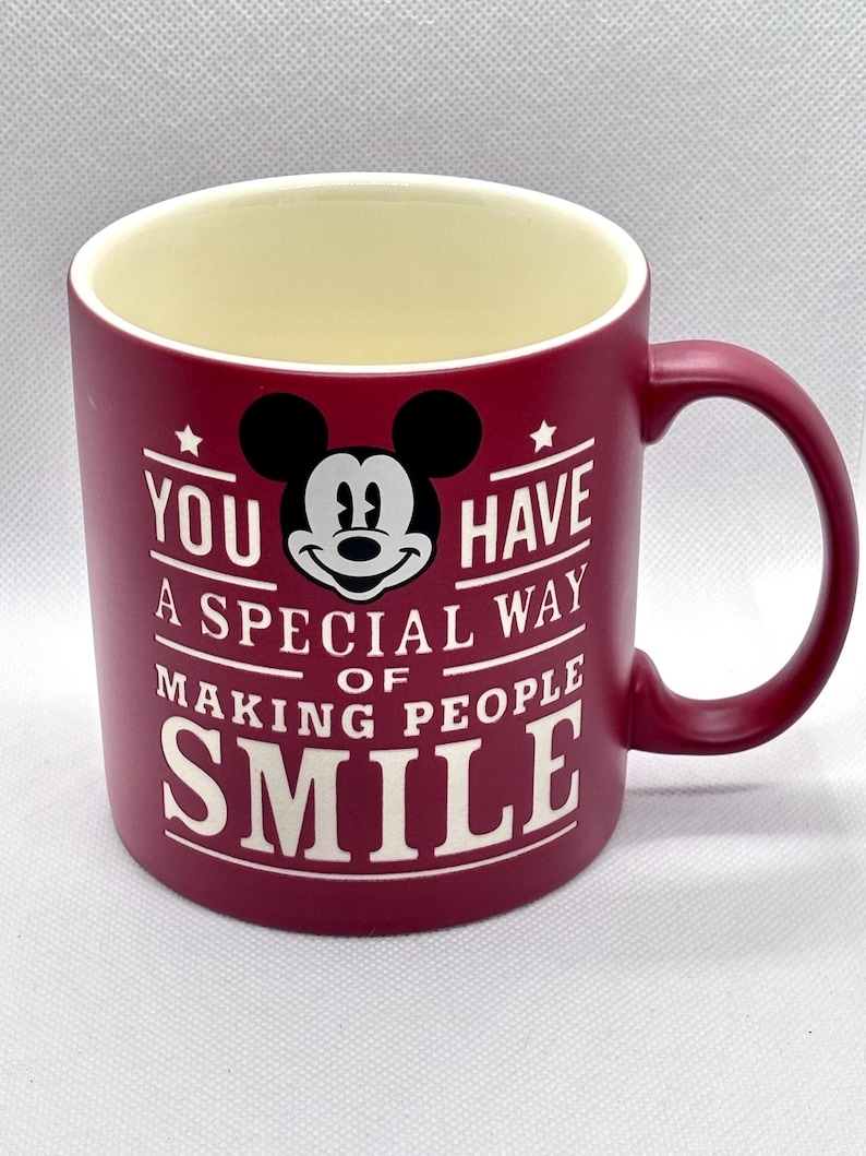Puede incluir: Una taza de cer&aacute;mica color burdeos mate con interior crema y asa en el lado derecho. El frente presenta un gr&aacute;fico en blanco y negro de la cabeza de Mickey Mouse sobre el texto blanco: YOU HAVE A SPECIAL WAY OF MAKING PEOPLE SMILE.