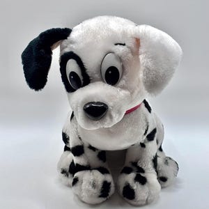 May include: A plush dalmatian dog toy with white fur and black spots. The stuffed animal features large plastic eyes, a black nose, and a pink collar around its neck. One ear is black while the other is white, and it is sitting in an upright position.