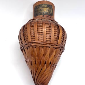 May include: A brown woven bamboo vessel with a tapered, teardrop-shaped body. The top features a smooth wooden neck section with a rectangular label printed with the text Souvenir of MIAMI, FLA in black ink against a dark background.