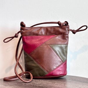 Puede incluir: Un bolso tipo cubo con un diseño de parches en paneles de cuero marrón, verde oliva y magenta. El bolso tiene un borde superior marrón oscuro, un cierre de cordón con extremos anudados y una correa de hombro fina y a juego en color marrón.