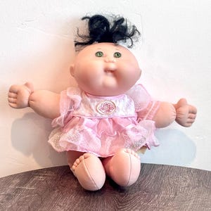 May include: A soft-bodied doll with a round face, green eyes, and black yarn hair styled in a top knot. The doll wears a light pink dress featuring ruffled lace sleeves, a polka-dot skirt overlay, and a circular emblem on the chest with CPK text.