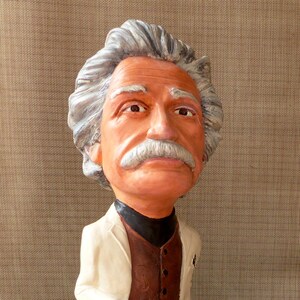 May include: A figurine of a person with white hair and a brown mustache wearing a white jacket, brown vest, and black pants. The figurine is standing on a brown base with the word 'Emc' inscribed on it.