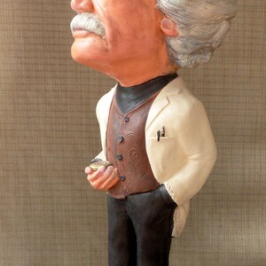 May include: A white ceramic figurine of a person with gray hair, wearing a white jacket, brown vest, and black pants. The figurine is standing on a brown base with a clock face and the letters 'E=mc2'.