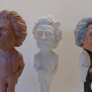 May include: Three ceramic figurines of men with different hair styles and colors. The figurine on the left is brown, the figurine in the middle is white, and the figurine on the right is brown and white.