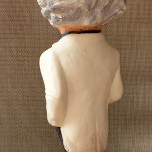 May include: A white ceramic figurine of a person with a white, textured hair style. The figurine is wearing a white jacket and black pants. The figurine is standing on a brown base with a design on it.